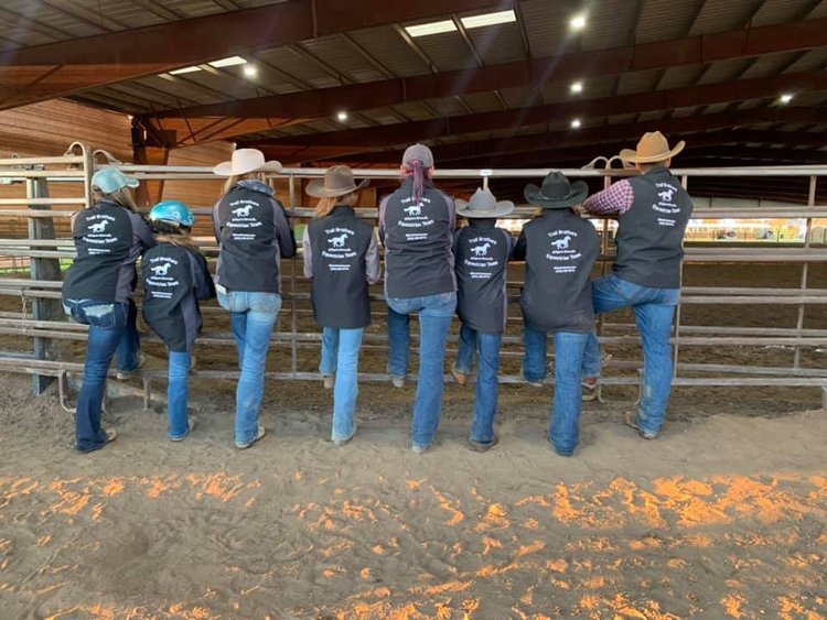 A line of riders in matching jackets standing together at the arena rail at Ocean View Stables, backlit by low sun