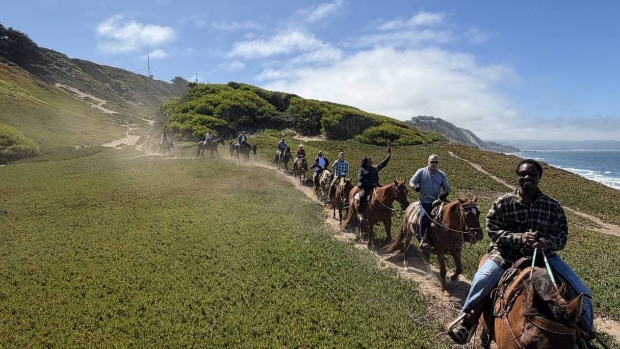 Riders climbing a green ridge trail with the coastline behind them above Ocean View Stables in Daly City, California